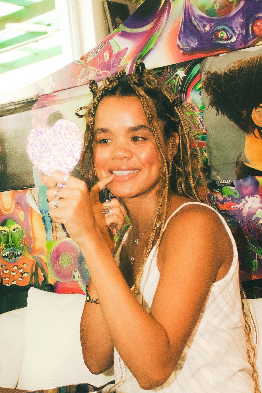 A woman in a white tank top  sits at a table in a vibrant, graffiti-covered room, admiring her tooth gem through a pink heart-shaped mirror.